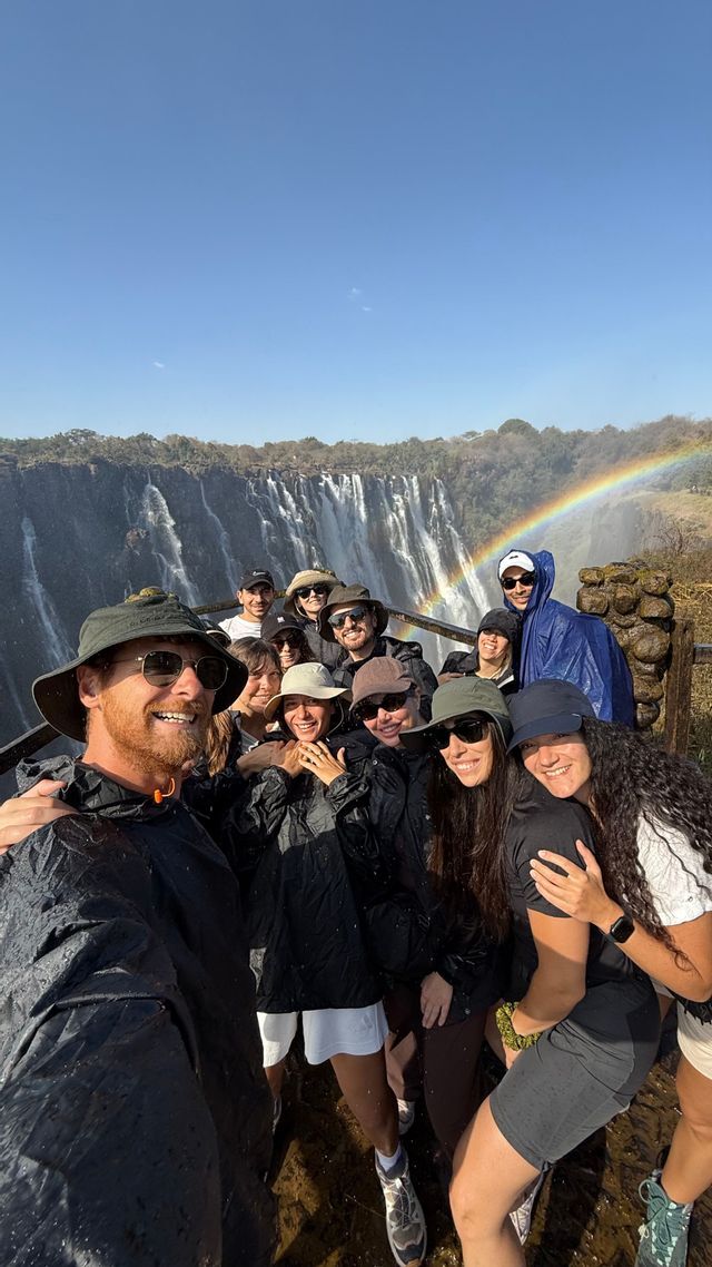 Un gruppo WeRoad si fa un selfie con attrezzatura antipioggia di fronte a una grande cascata con un arcobaleno nel cielo.