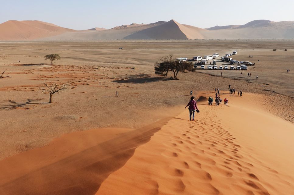 A WeRoad group trip walks up the ridge of a large orange sand dune in a vast desert landscape.