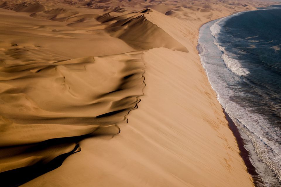 Una vista aerea di grandi dune di sabbia ondulate che incontrano direttamente la costa di un oceano blu scuro con onde bianche.