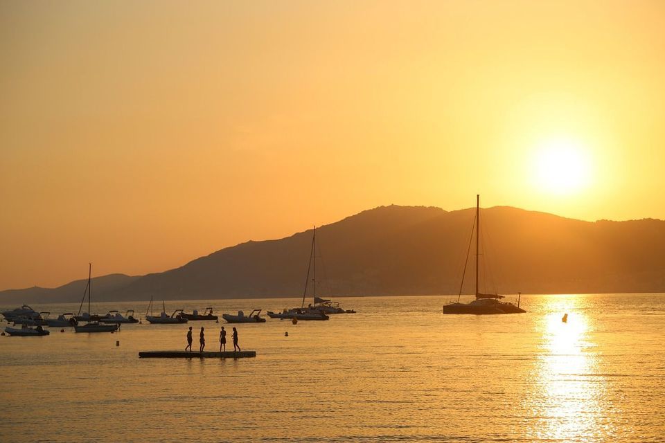 Silhouettes d'un groupe WeRoad en voyage sur un ponton flottant dans une baie, avec des bateaux, au coucher du soleil derrière les collines.