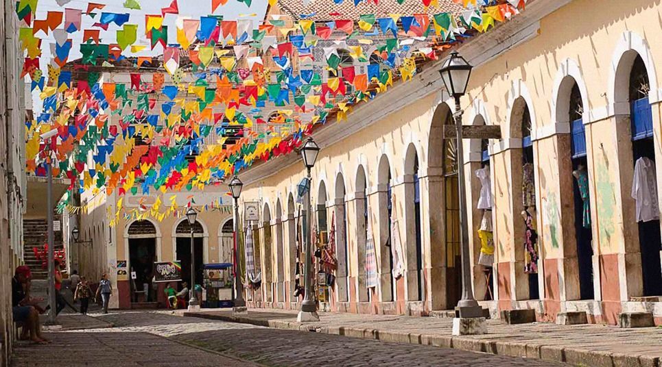 Colorful festival flags hang over a cobblestone street lined with yellow buildings with arched colonnades.