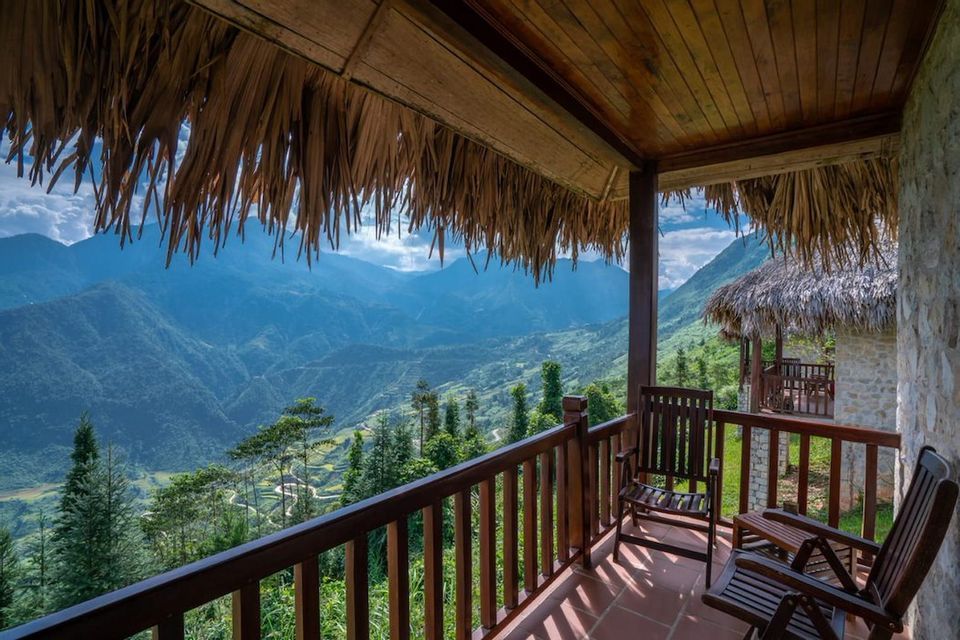 Vista da un balcone con tetto di paglia e due sedie in legno che si affaccia su una vasta valle di montagna verde in una giornata di sole.