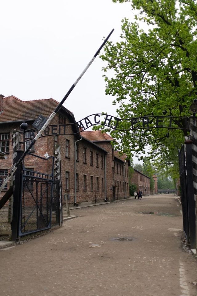 El letrero 'Arbeit Macht Frei' sobre la puerta de entrada principal de un antiguo campo de concentración, con barracones de ladrillo bordeando un camino de tierra.
