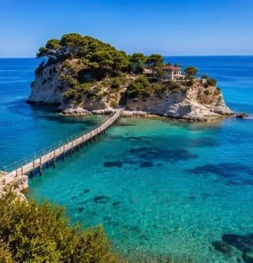 Un ponte di legno si estende su acque cristalline color turchese fino a una piccola isola rocciosa, coperta di alberi, sotto un cielo azzurro brillante.