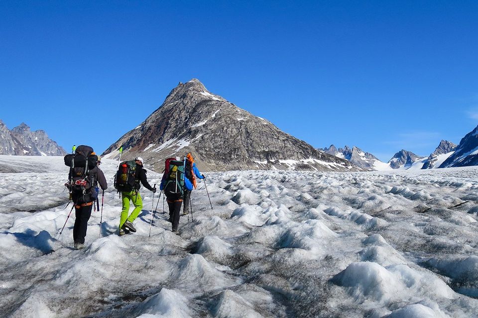 Un viaggio di gruppo WeRoad con tre escursionisti che, muniti di zaini, fanno trekking su un vasto ghiacciaio ondulato, diretti verso una montagna rocciosa sotto un cielo azzurro e limpido.