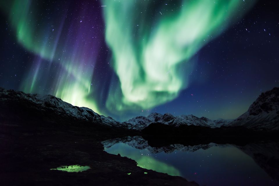 L'aurora boreale verde e viola brilla nel cielo notturno, riflettendosi su un lago calmo e montagne innevate.