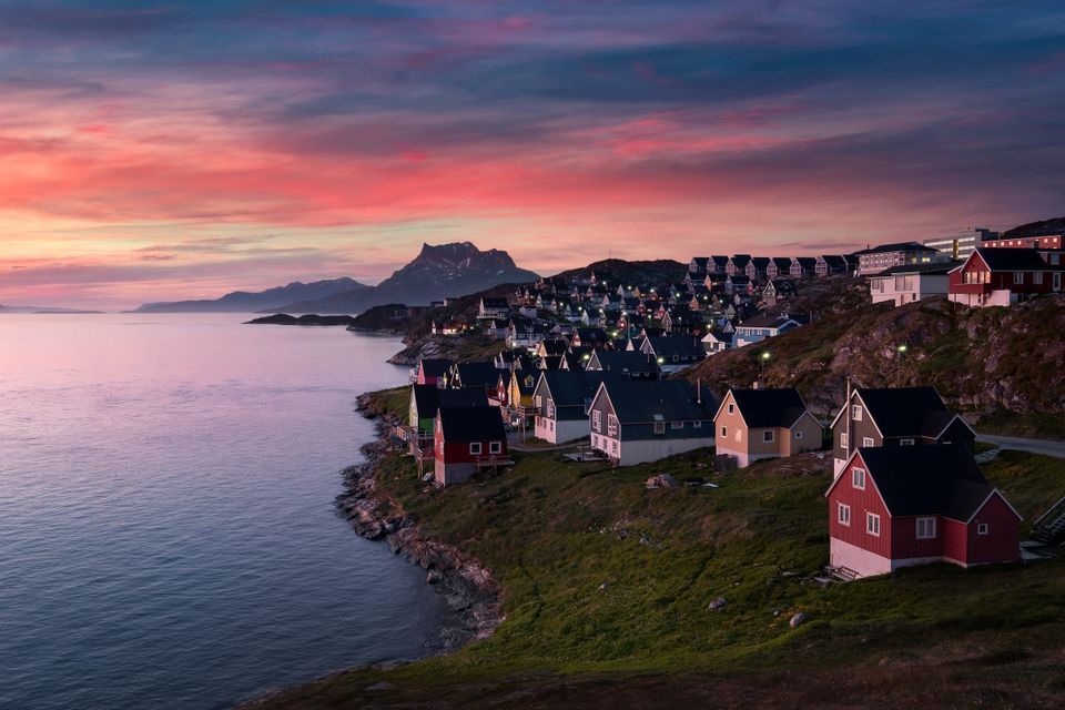 Un borgo costiero con case colorate è illuminato lungo una costa rocciosa al tramonto, con un cielo vibrante di rosa e viola.