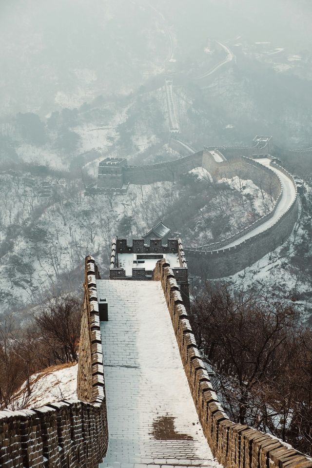 Una veduta dall'alto di una sezione innevata della Grande Muraglia cinese che si snoda attraverso un paesaggio nebbioso e boscoso.