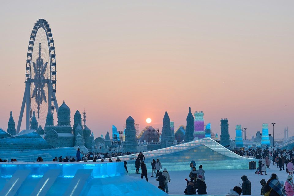 Un'ampia vista di un festival invernale con persone tra grandi sculture di ghiaccio e una ruota panoramica contro un cielo al tramonto.