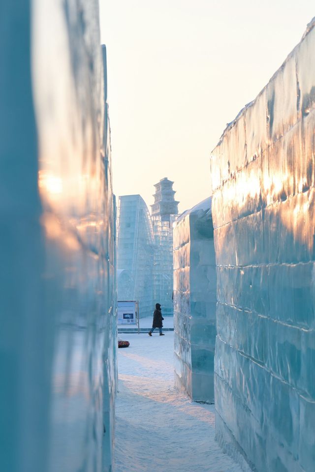Una persona cammina su un sentiero innevato tra imponenti pareti di ghiaccio, con una pagoda di ghiaccio visibile in lontananza al tramonto.