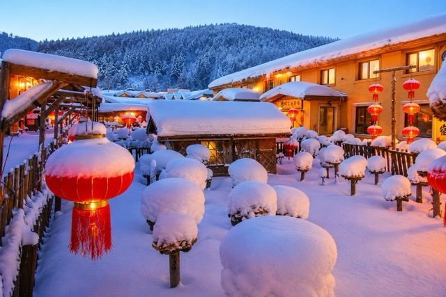 Un villaggio innevato al crepuscolo, con lanterne rosse accese, edifici illuminati da una luce calda e lo sfondo di una foresta imbiancata.