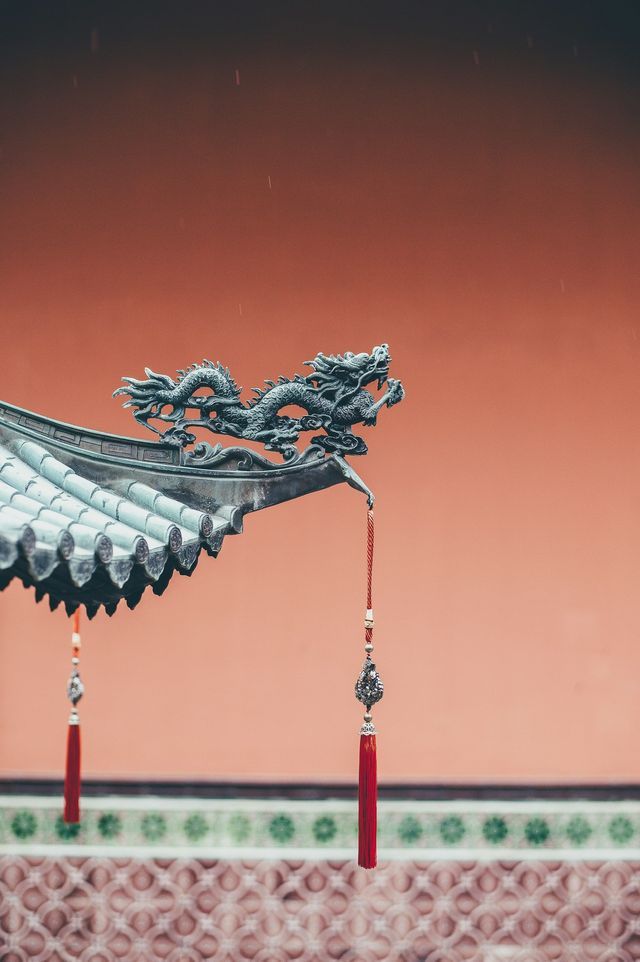 Un primo piano di una scultura di drago in metallo sulla grondaia di un tetto in tegole, con una nappa rossa pendente contro un muro rosso.