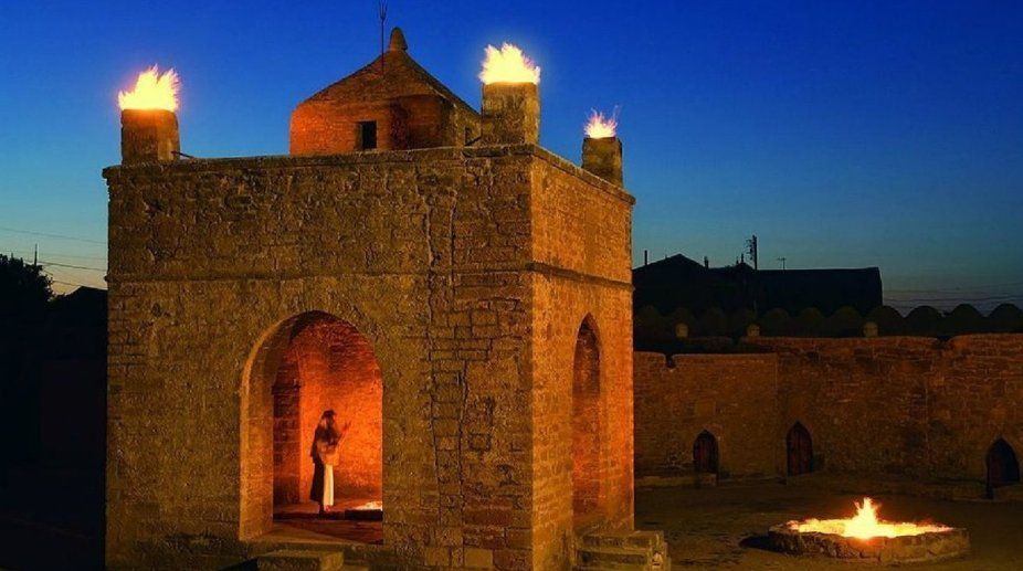 Ein Steintempel mit Feuern auf dem Dach und einer Feuerstelle im Innenhof bei Dämmerung, mit einer Person, die in einem Torbogen steht.