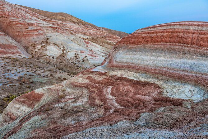 Schichten aus rotem, weißem und grauem Sediment bilden sanfte, gestreifte Hügel unter einem hellblauen Himmel.