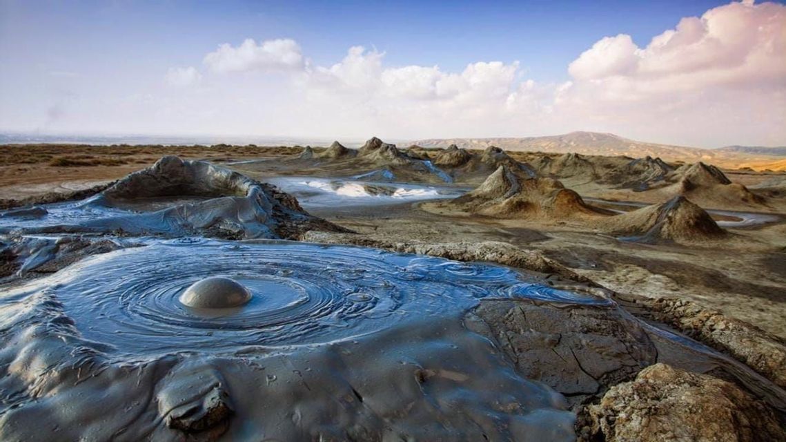 Ein blubbernder Schlammvulkan erzeugt konzentrische Wellen in einer ariden Landschaft mit zahlreichen kleineren Schlammhügeln unter bewölktem Himmel.
