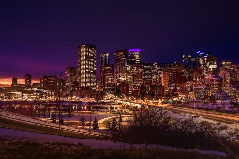 Une ligne d'horizon urbaine de gratte-ciel illuminés, sous un ciel de crépuscule pourpre, avec un sol enneigé et un pont éclairé au premier plan.