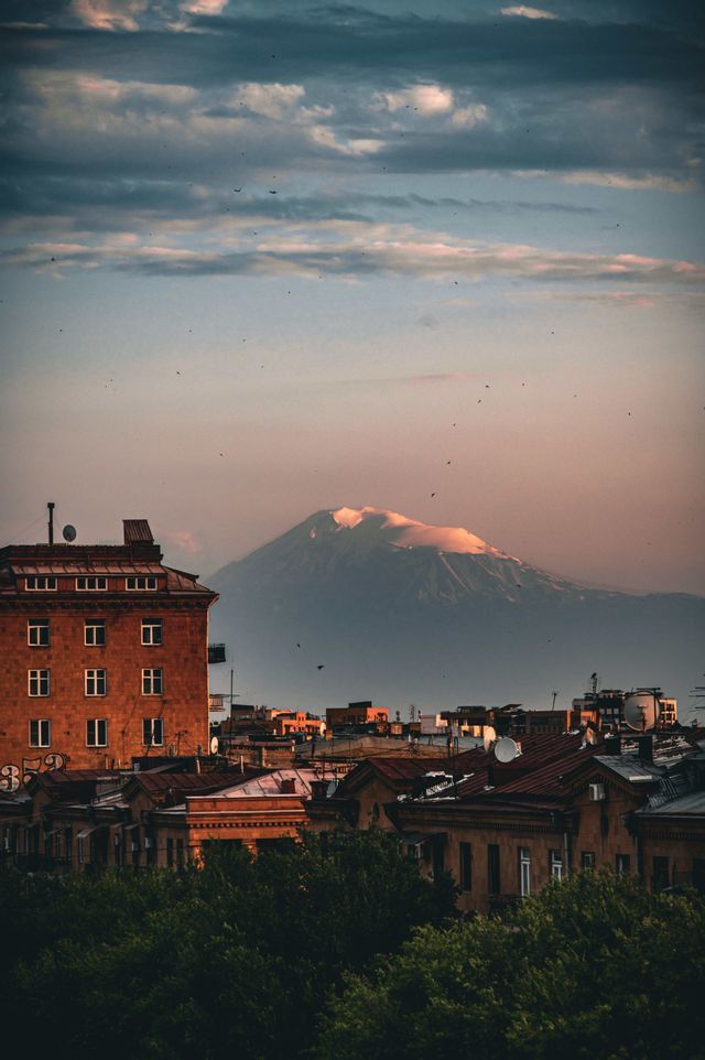 Une grande montagne enneigée domine les toits de la ville et la cime des arbres verts sous un ciel de coucher de soleil nuageux.