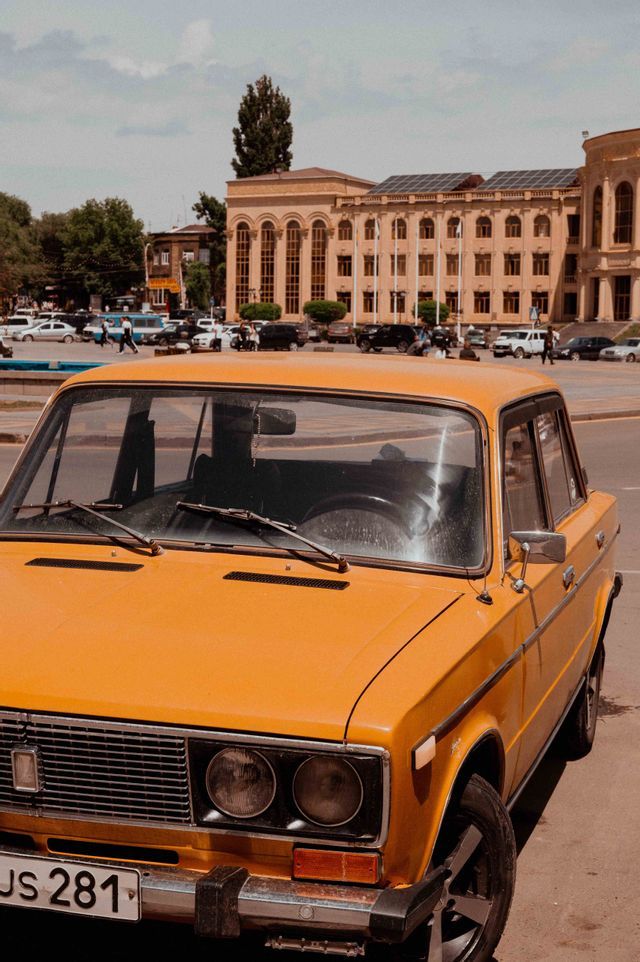 Une voiture ancienne jaune garée sur une place de ville devant un grand bâtiment classique avec des gens en arrière-plan.