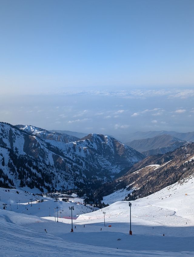 Vue plongeante d'une vallée de montagne enneigée avec des skieurs sur une piste sous un ciel bleu dégagé.