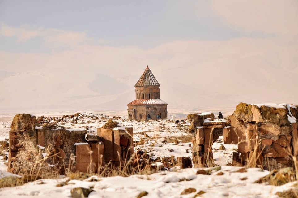 Une ancienne église en pierre au toit conique se dresse dans un vaste paysage enneigé, vue de derrière des ruines délabrées.