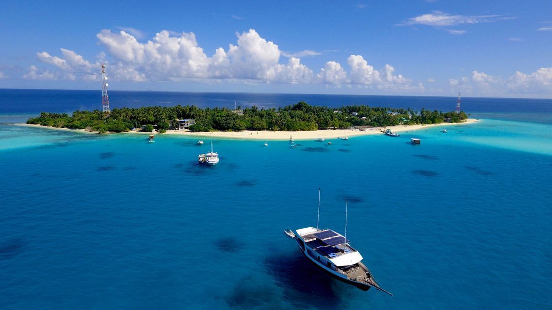 An aerial view of several boats anchored in turquoise water around a tropical island with a sandy beach and palm trees.