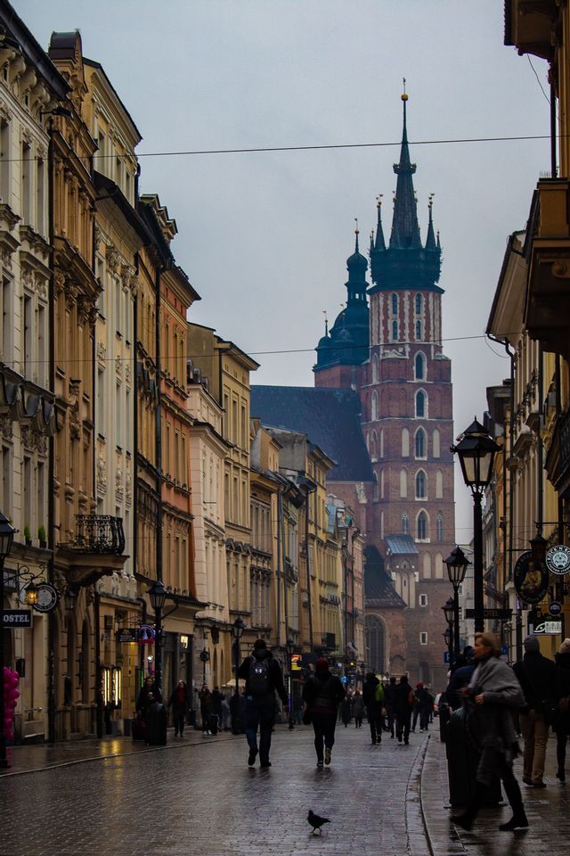 Des gens marchent dans une rue pavée et humide, bordée de bâtiments historiques, menant à une grande église en briques avec une tour proéminente.
