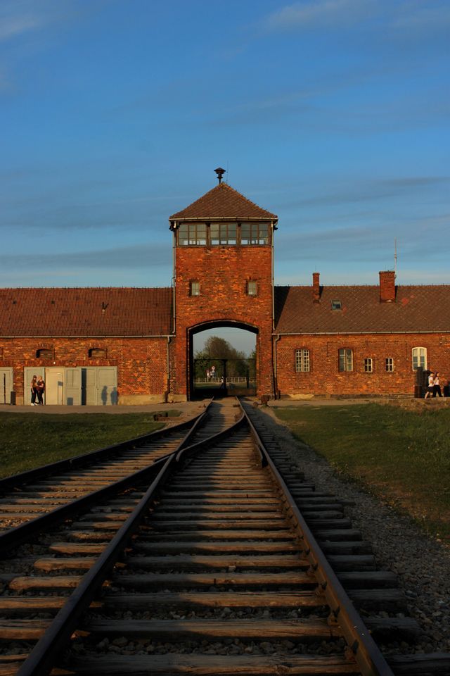 Des rails de chemin de fer mènent vers l'entrée voûtée d'un bâtiment en briques rouges, doté d'une tour de guet, sous un ciel bleu au crépuscule.