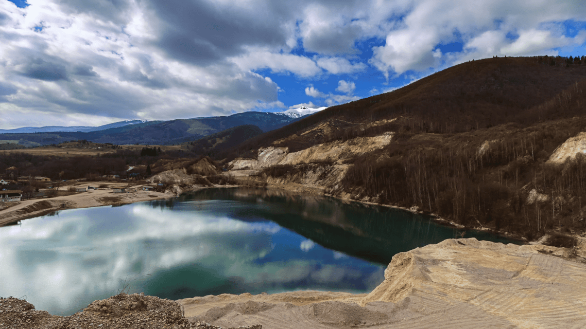 Un lago calmo in una cava riflette un cielo azzurro nuvoloso, con colline brulle e lontane montagne innevate sullo sfondo.