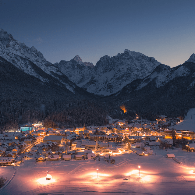 Un villaggio alpino illuminato brilla in una valle innevata ai piedi di imponenti montagne sotto un cielo crepuscolare.