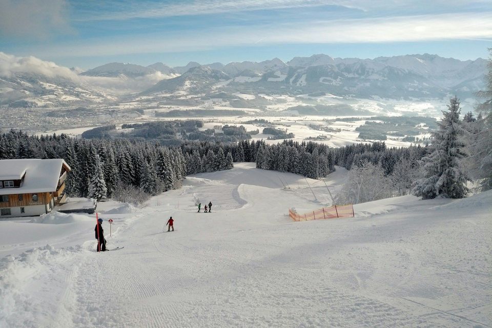 Sciatori su un'ampia pista innevata vicino a uno chalet, con vista su una pineta e una vasta valle di montagna.