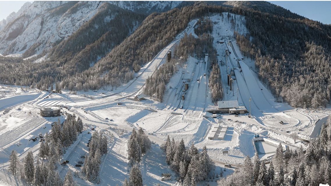 Una vista aerea di un complesso per il salto con gli sci innevato, integrato in un versante montuoso boscoso.