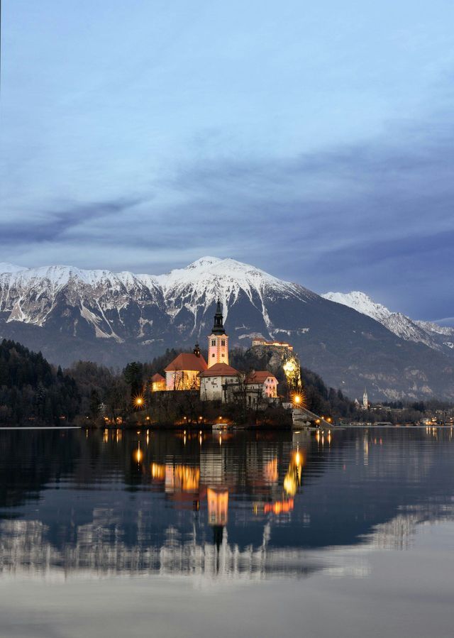 Una chiesa illuminata su una piccola isola si riflette nelle acque calme di un lago al tramonto, con montagne innevate sullo sfondo.