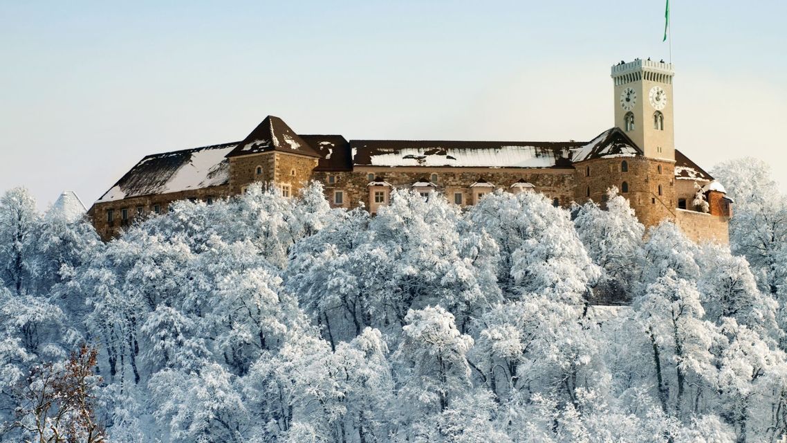 Un castello in pietra con una torre dell'orologio si erge sopra una fitta foresta di alberi completamente coperti di neve bianca.