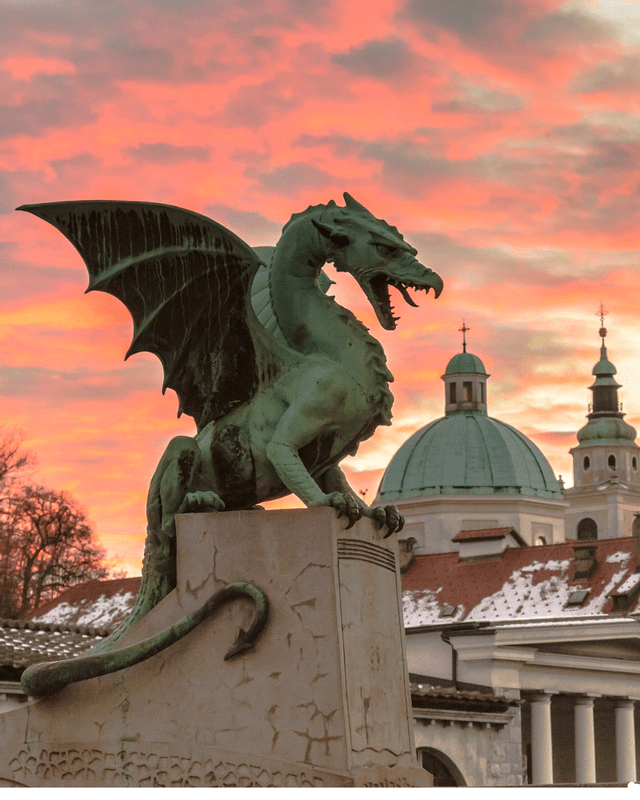 Una grande statua di drago alato in bronzo si erge su un piedistallo di pietra, stagliandosi contro un cielo colorato al tramonto su una città con cupole.
