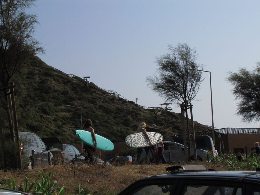 Un viaggio di gruppo WeRoad trasporta tavole da surf attraverso un parcheggio con una grande collina verde sullo sfondo.