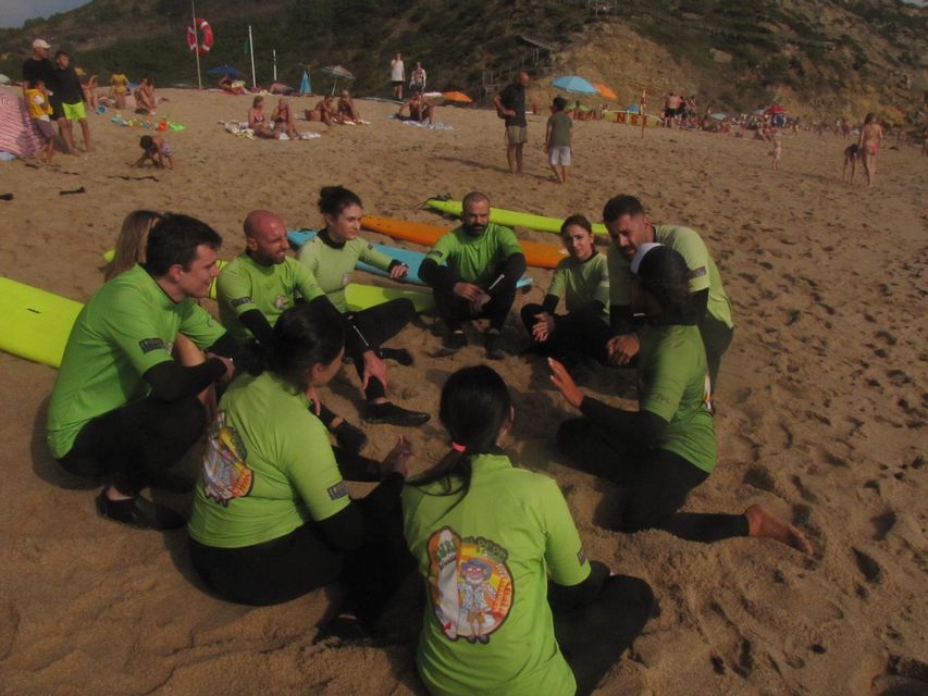 Un gruppo WeRoad, che indossa top verdi e mute abbinate, è seduto in cerchio su una spiaggia sabbiosa con le tavole da surf.