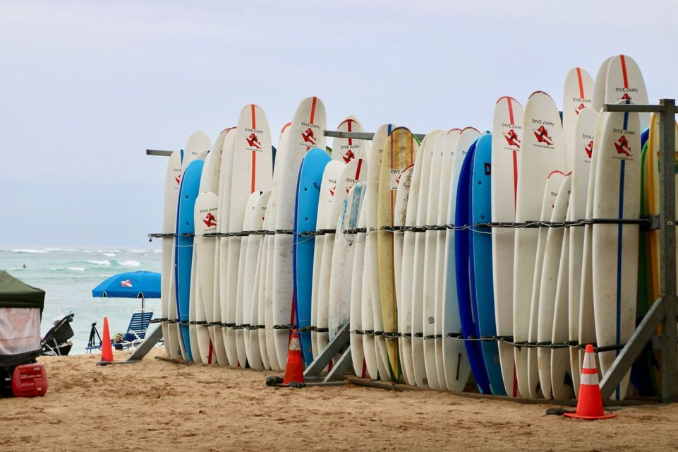 Una grande rastrelliera di tavole da surf bianche e blu si trova su una spiaggia sabbiosa, con le onde dell'oceano visibili sullo sfondo.