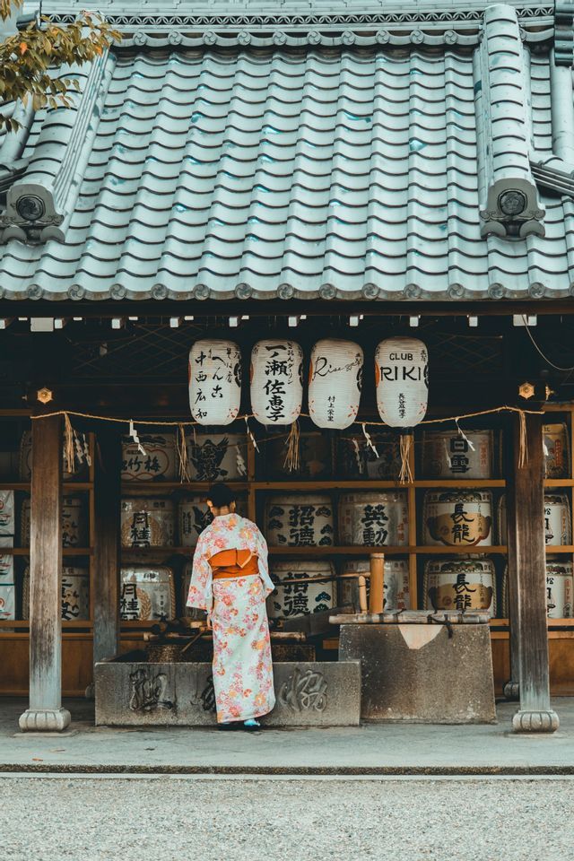 A person seen from behind wearing a floral kimono stands in front of a traditional Japanese temple with hanging lanterns.