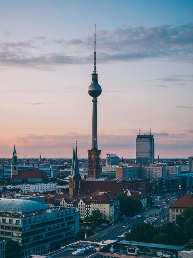 Veduta aerea di una città al tramonto, con un'imponente torre televisiva che si innalza sopra gli edifici e le chiese.