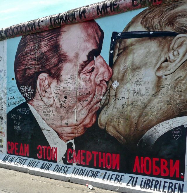 Un primo piano del murale 'Bacio Fraterno' sul Muro di Berlino, che raffigura due uomini che si baciano, coperto di graffiti.