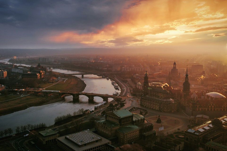 Veduta aerea di una città storica con un ampio fiume e un ponte ad arco, illuminata dalla calda luce di un tramonto dorato.