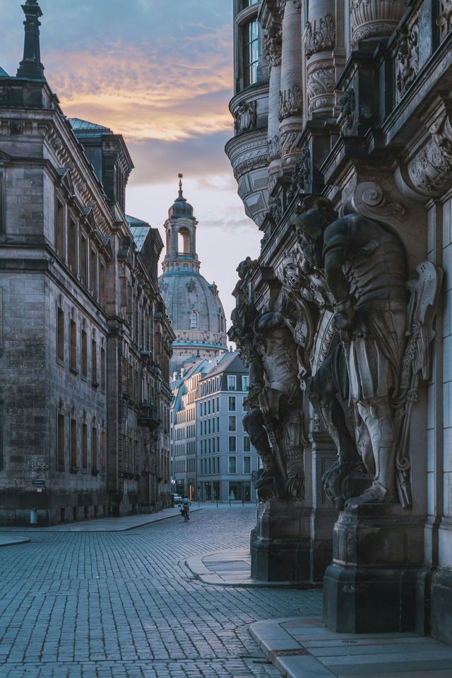 Scorcio di una strada acciottolata tra eleganti palazzi in pietra, con una grande cupola sullo sfondo di un cielo al tramonto colorato.