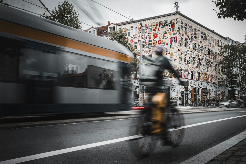 Un ciclista su una strada accanto a un tram in transito, entrambi ripresi con effetto mosso davanti a un edificio coperto di arte colorata.