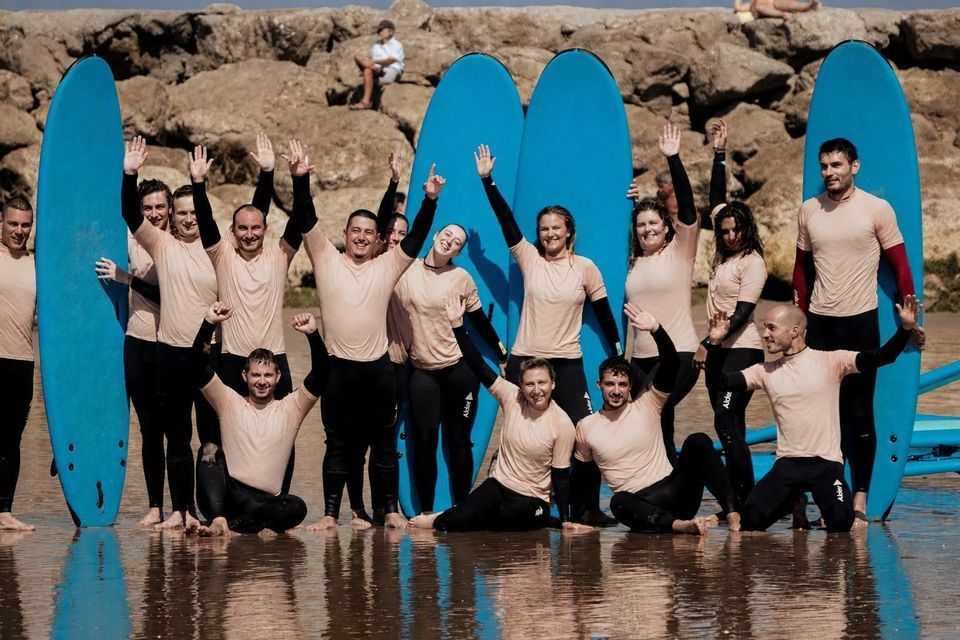 Un gruppo WeRoad in mute da sub, in posa per una foto su una spiaggia, sorridenti e che salutano con le loro tavole da surf blu.