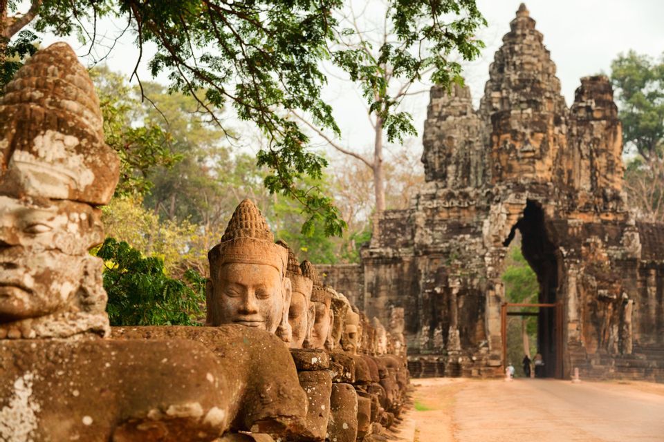 A row of weathered stone statues with carved faces lines a road leading to a large, ornate temple gate with trees overhead.
