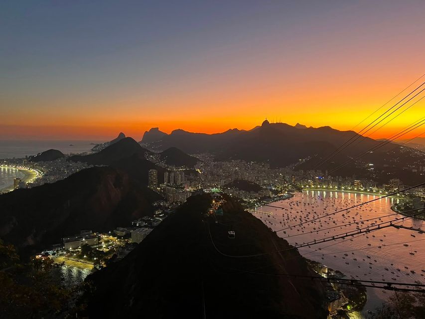 Vista dall'alto di una città costiera illuminata al tramonto, con montagne in silhouette e una baia piena di barche.