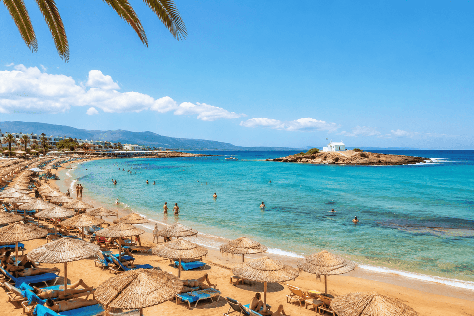 Una veduta dall'alto di una spiaggia sabbiosa affollata con ombrelloni di paglia, persone che nuotano nell'acqua turchese e una piccola isola con una cappella.
