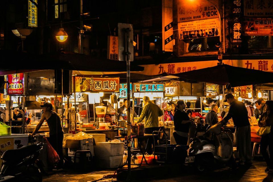 Un vivace mercato di strada notturno con persone riunite attorno a bancarelle di cibo illuminate e insegne in caratteri asiatici.