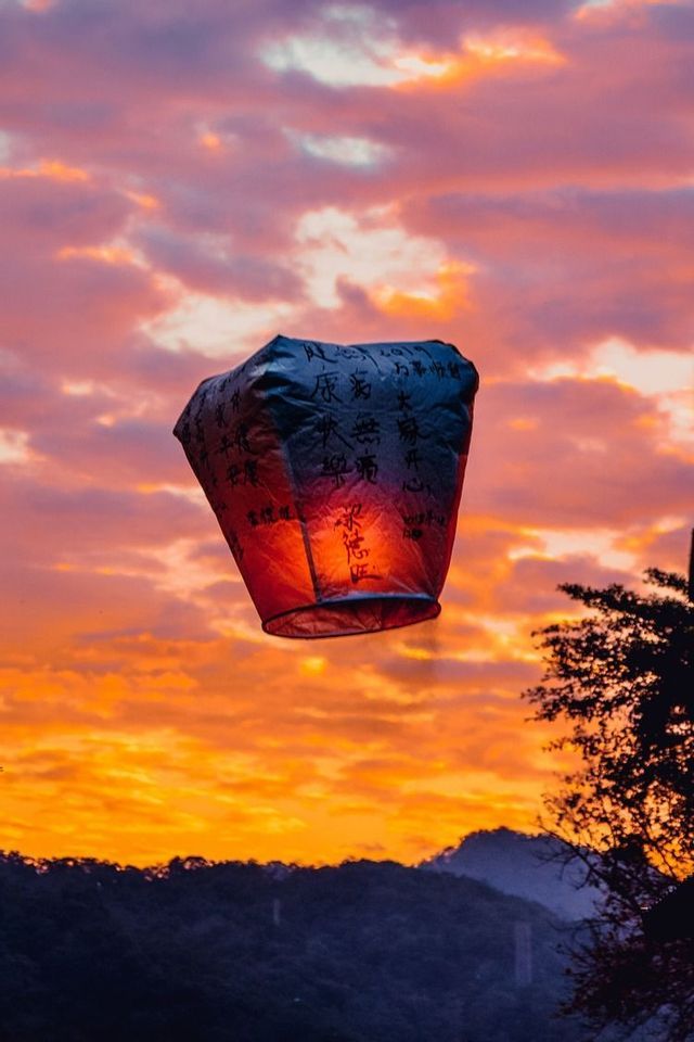 Una lanterna di carta illuminata con calligrafia fluttua in un cielo nuvoloso, arancione e viola al tramonto, sopra colline in silhouette.