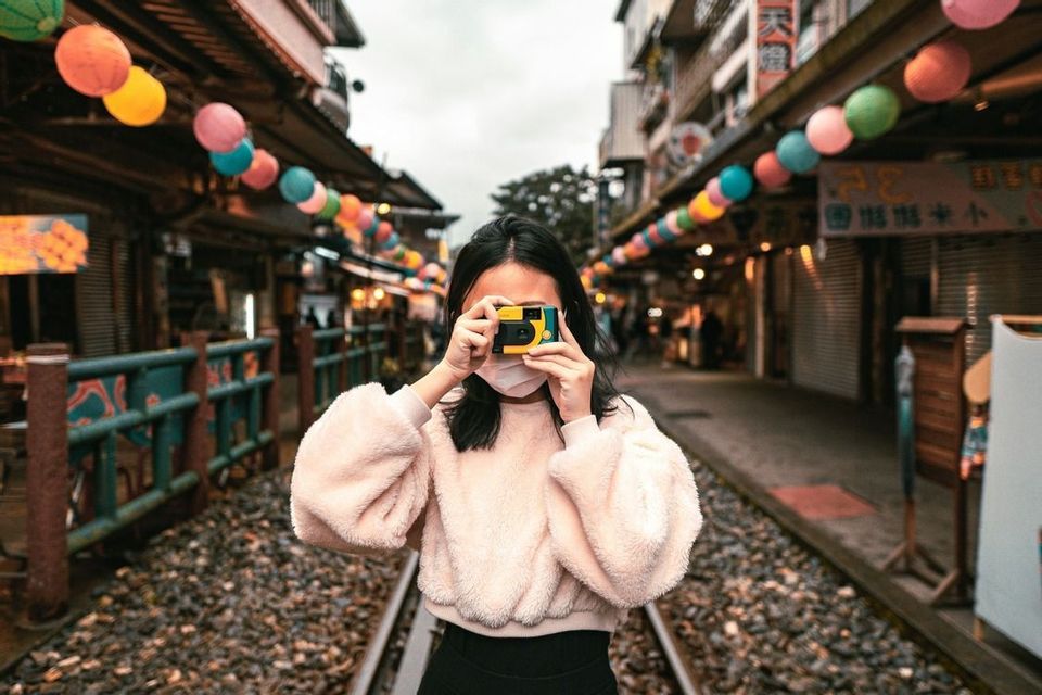 Una donna con la mascherina scatta una foto con una macchina fotografica, stando sui binari del treno in una strada di mercato con lanterne colorate.